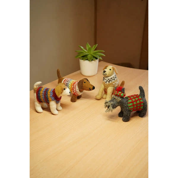 Felt dog figurines wearing colorful sweaters, arranged on a wooden surface with a green potted plant in the background.
