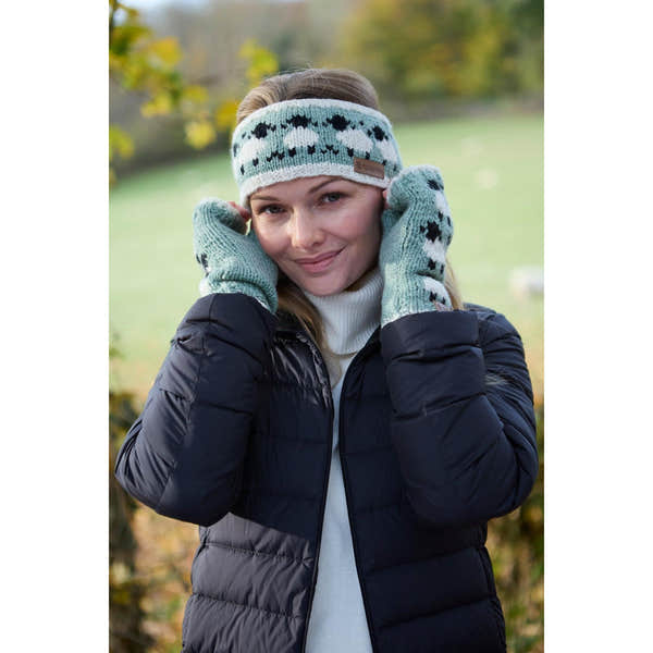 Pachamama Merino sheep handwarmer and headband set in green with sheep pattern, worn by a woman outdoors in fall.
