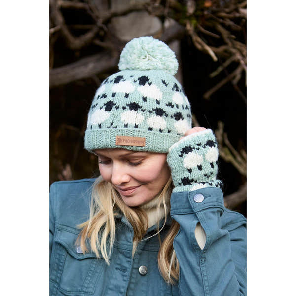 Woman wearing green Pachamama Merino sheep knit beanie and mittens, outdoor winter accessories, with pom-pom and sheep pattern.