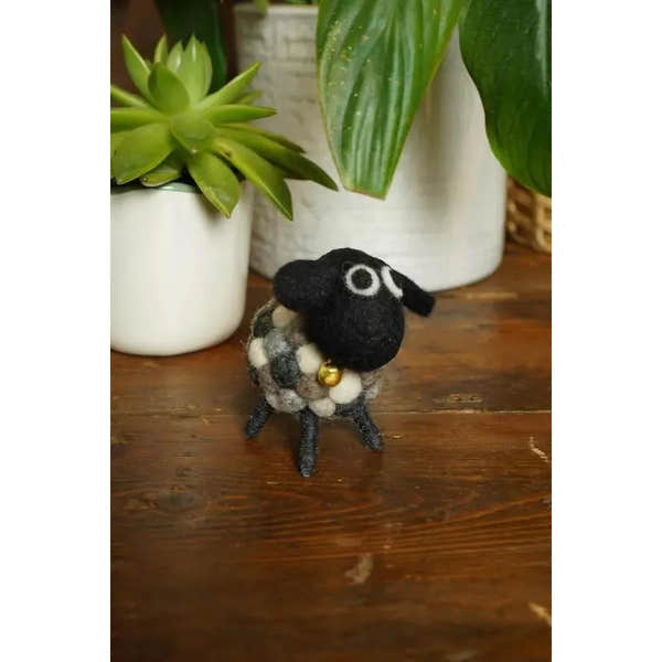 Pachamama Mini Helen felted sheep figurine with black head, white eyes, and checkered wool body, placed on a wooden surface near potted plants.