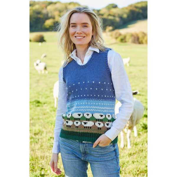 Pachamama Snowy Sheep knit tank top worn over a white shirt, featuring a colorful sheep pattern, outdoors in a green field.