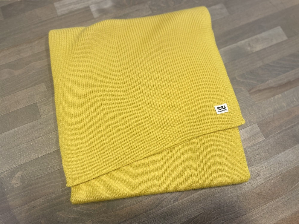 ROKA London Hyde Corn Scarf in bright yellow, knitted fabric, folded on wooden surface, with ROKA logo tag.