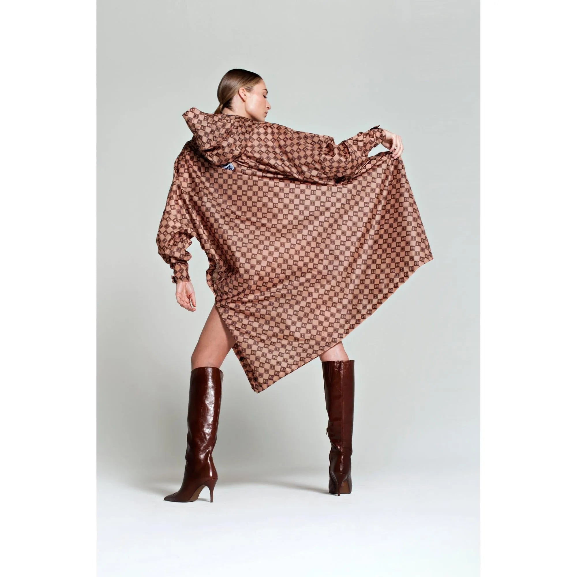 Rainkiss Damier rain poncho with checkered brown pattern, oversized design, and hood, styled with tall brown boots.