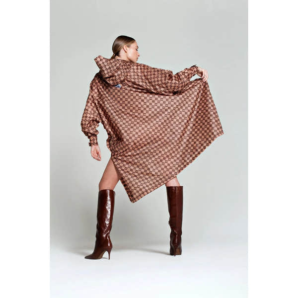 Rainkiss Damier rain poncho with checkered brown pattern, oversized design, and hood, styled with tall brown boots.