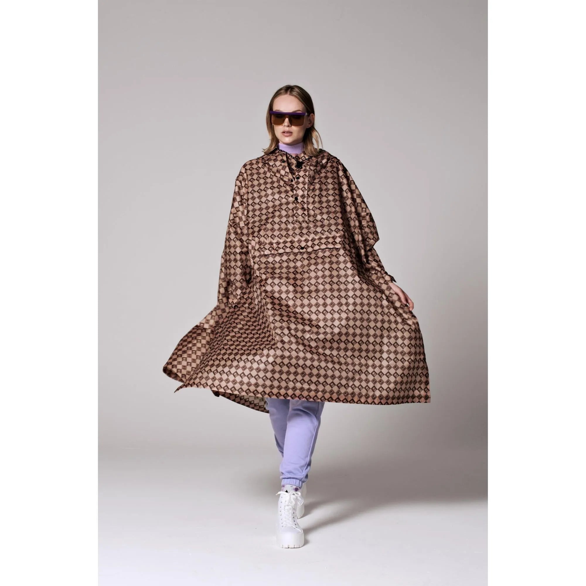 Rainkiss Damier rain poncho with a brown checkered pattern, oversized fit, and hood, styled with light purple pants and white boots.