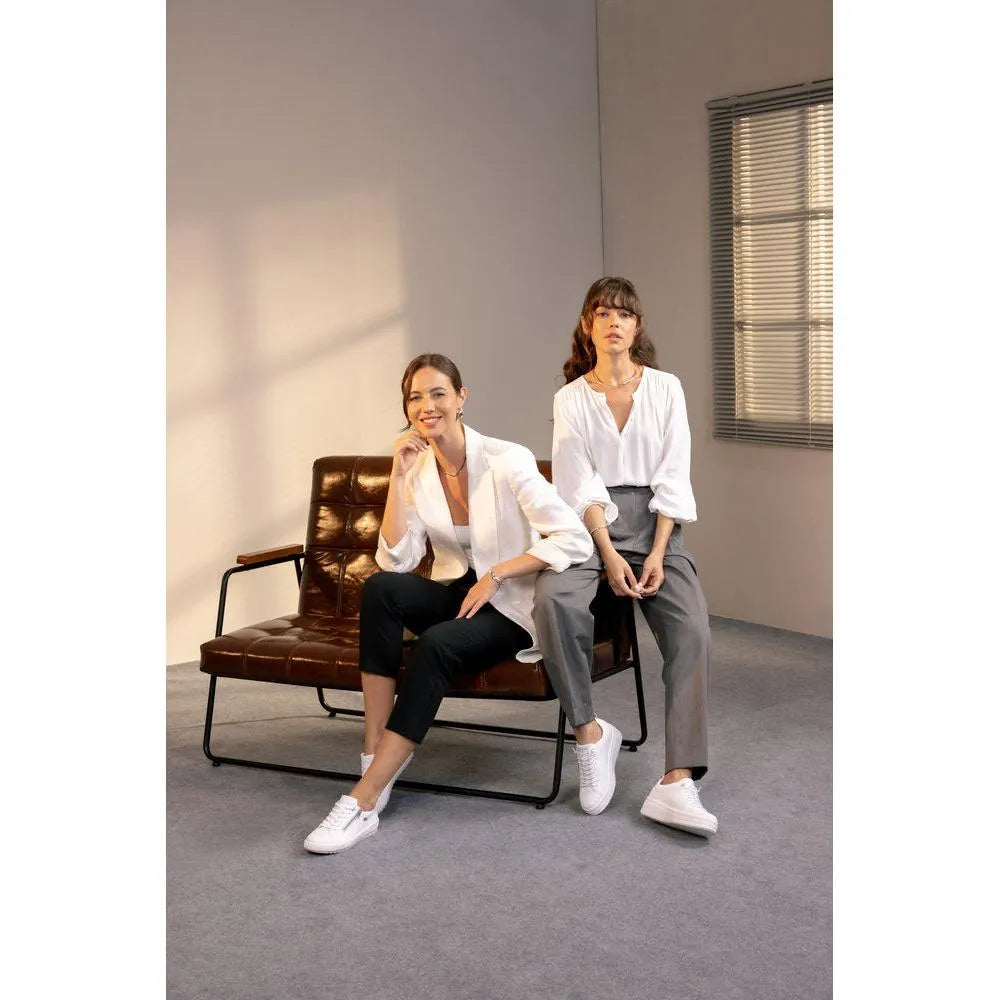 Women wearing white leather lace-up shoes, sitting on a leather sofa in a modern room with gray walls and window blinds.