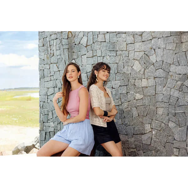Two women posing outdoors against a stone wall, showcasing fashion styles with natural landscape in background.