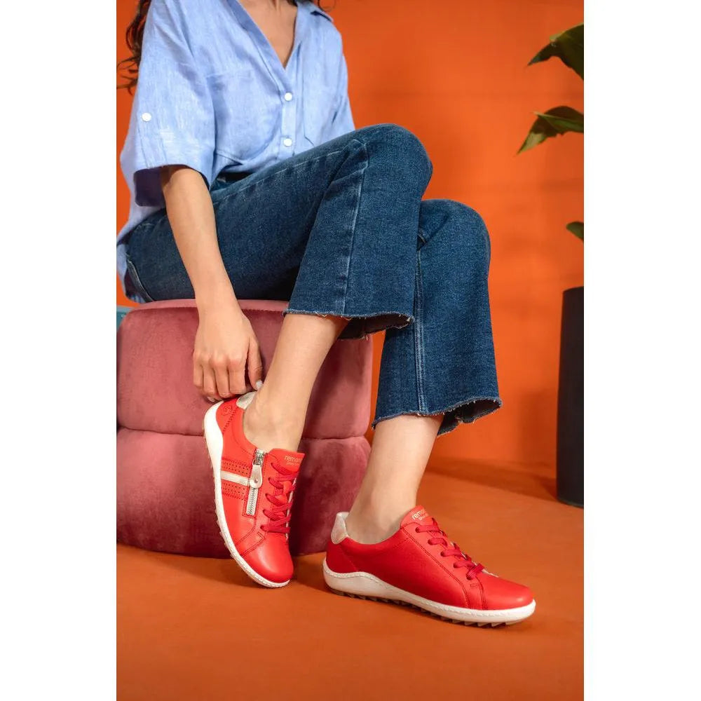 Woman wearing red leather Remonte R1432-34 lace-up shoes with zip detail, denim pants, and a blue shirt against an orange background.