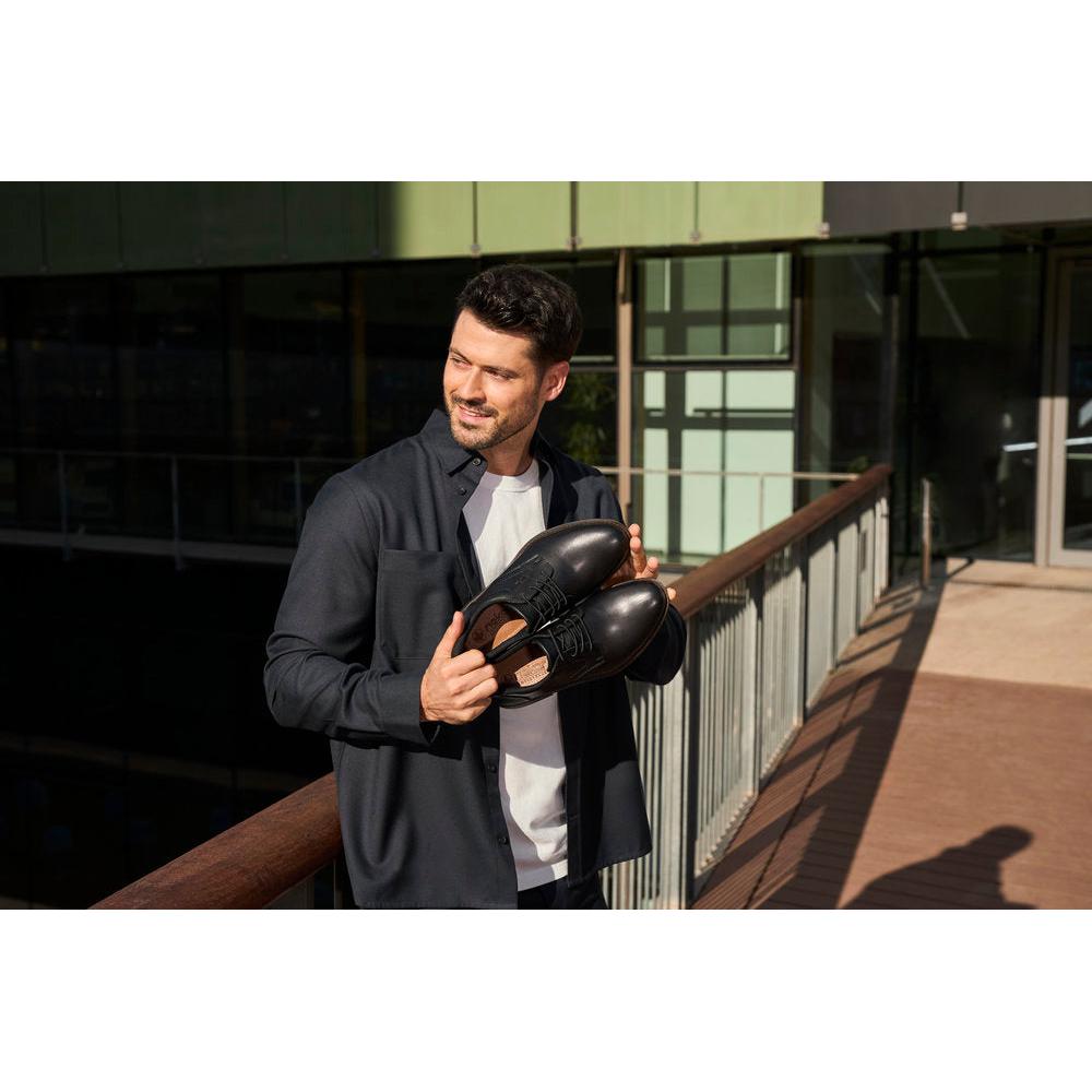 Man holding black leather slip-on shoes, stylish men's wide-fit dress shoes, outdoor urban setting.