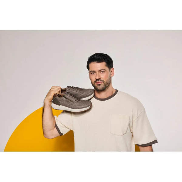 Man in beige shirt holding brown zip and lace shoes over shoulder, neutral background, yellow semi-circle design.