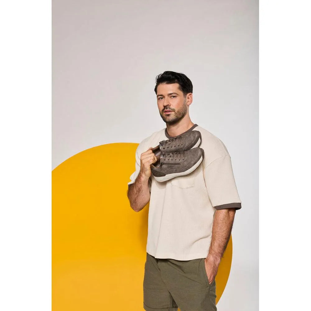 Man holding brown zip and lace shoes, wearing a beige shirt and olive pants, against a white background with yellow circle.