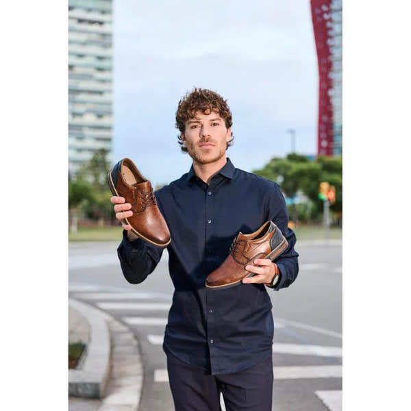 Man holding a pair of brown leather lace-up shoes outdoors in an urban setting.