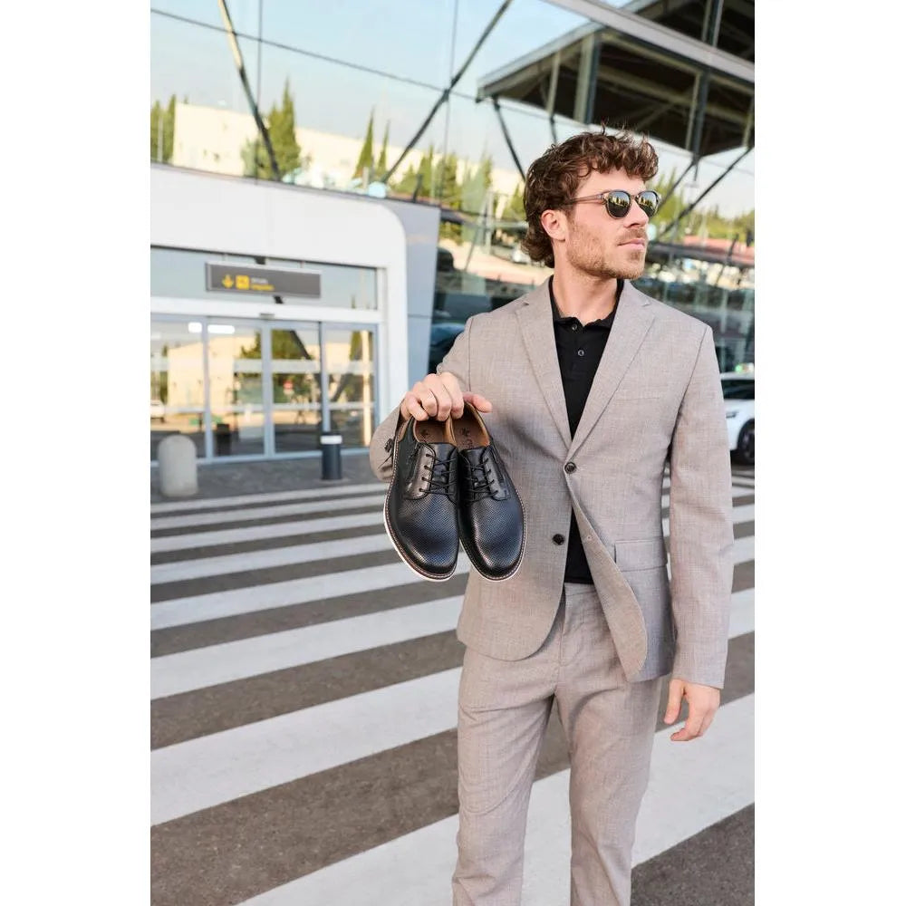 Man in gray suit holding black leather zip and lace shoes, standing outdoors near glass building.