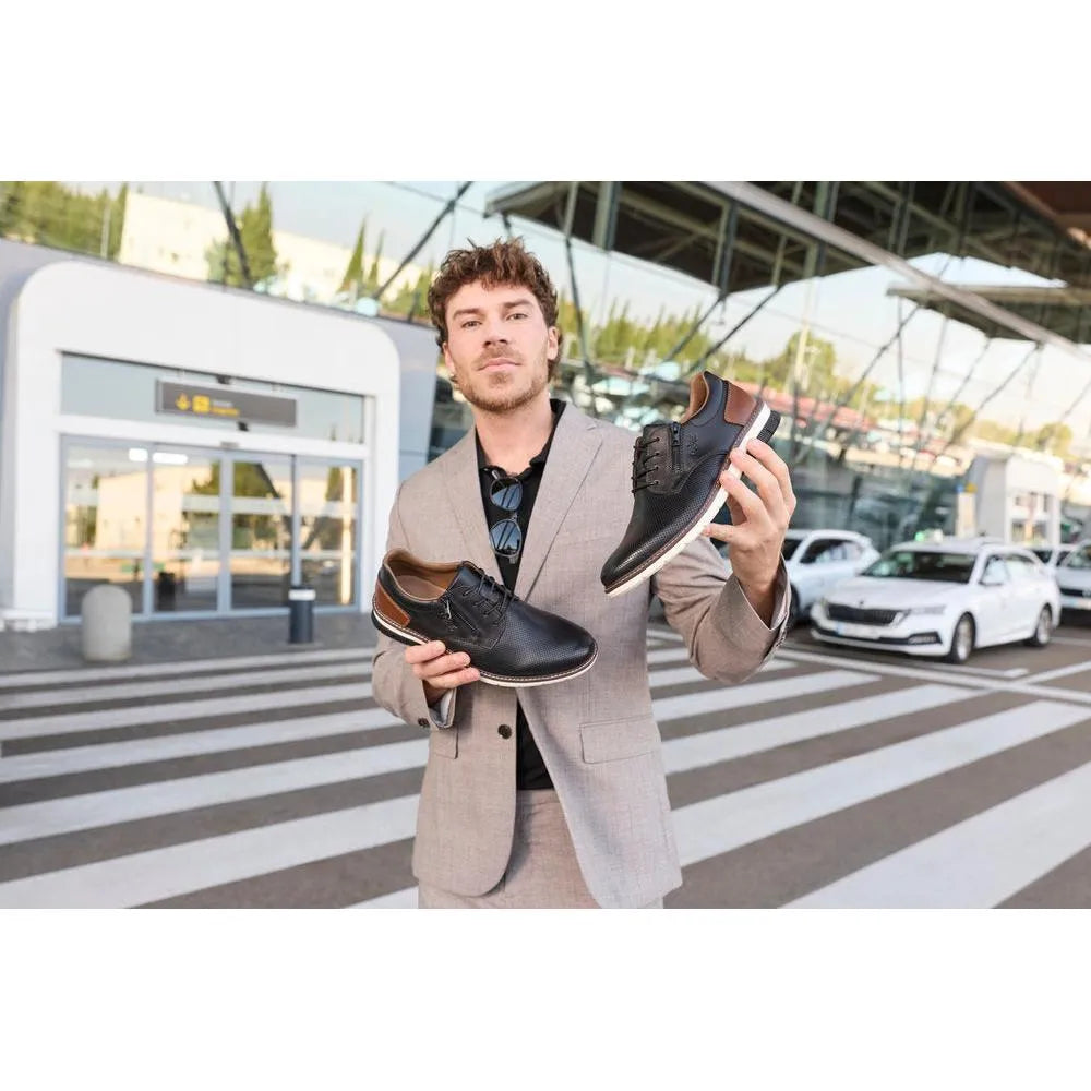 Man holding black leather zip and lace shoes, styled with brown accents, outside modern building and cars.