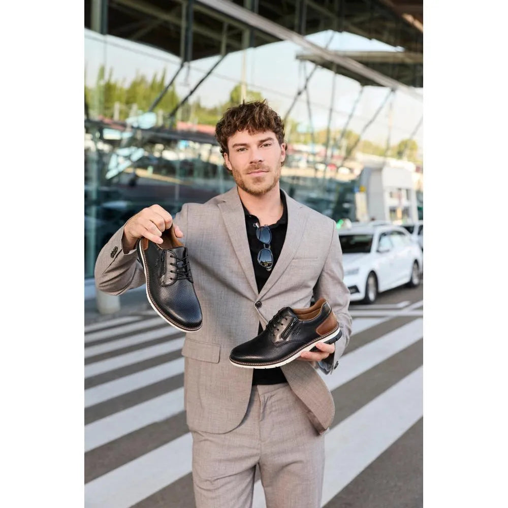 Man in a suit holding black leather dress shoes with lace and zip details, brown accents, and white soles, outdoors.
