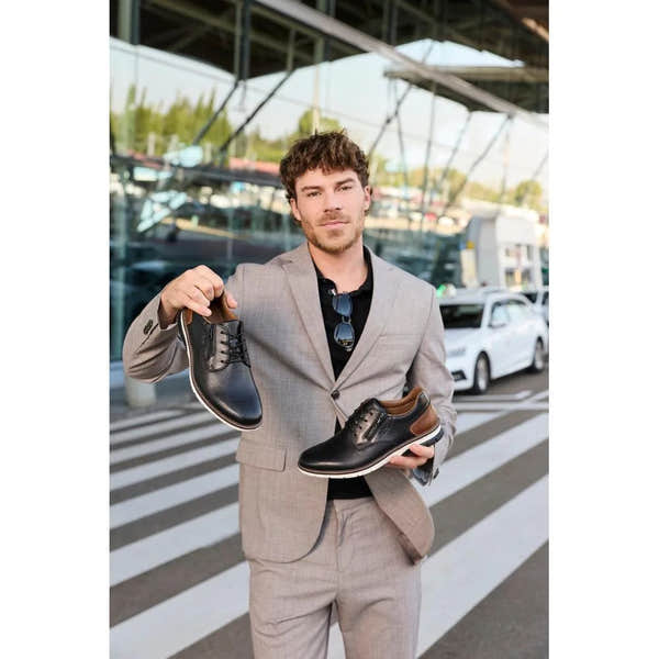 Man in a suit holding black leather dress shoes with lace and zip details, brown accents, and white soles, outdoors.