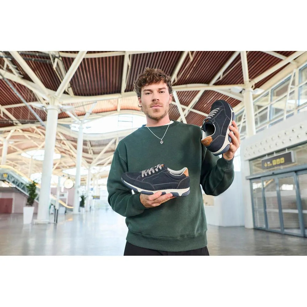 Man holding black and navy leather lace-up shoes with tan accents in a modern train station.