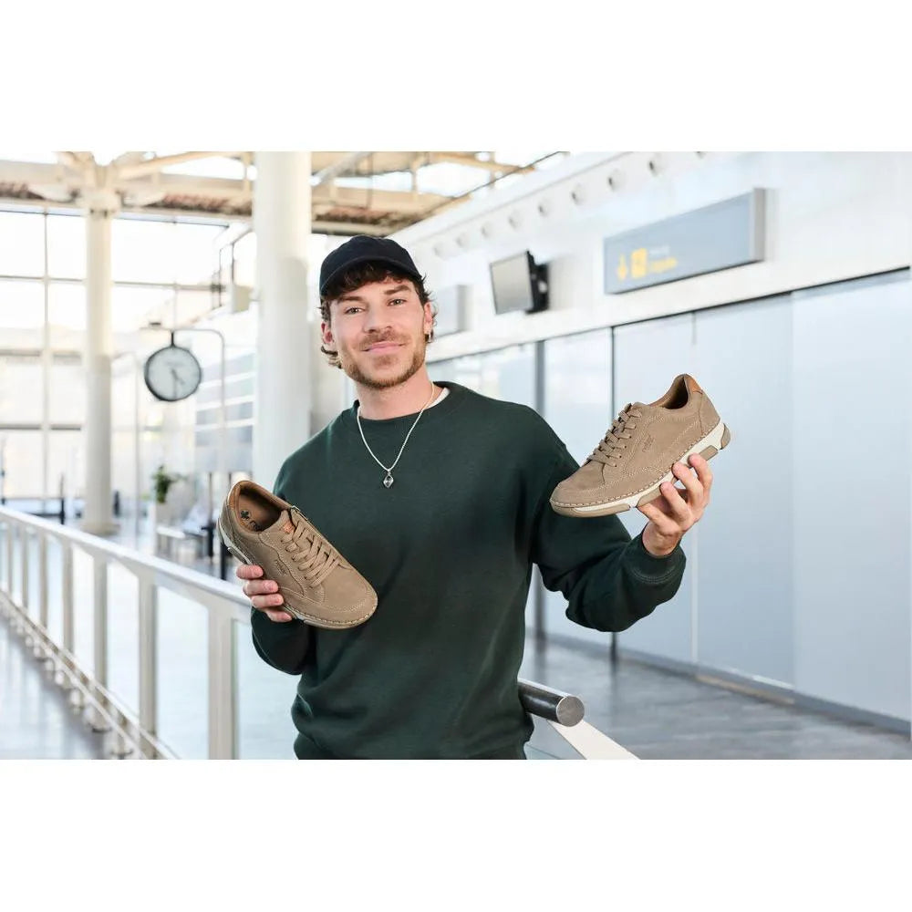 Man holding tan leather lace-up shoes, wide fit, casual style, in an airport terminal.
