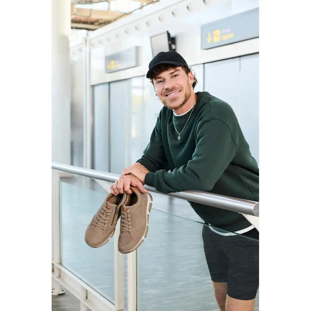 Man holding tan leather lace-up shoes, wearing a dark green sweatshirt, black shorts, and a cap at an airport.