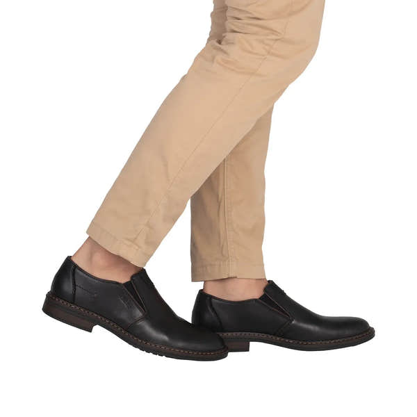 Black leather men's wide elasticated slip-on shoes with stitching detail, paired with beige trousers.