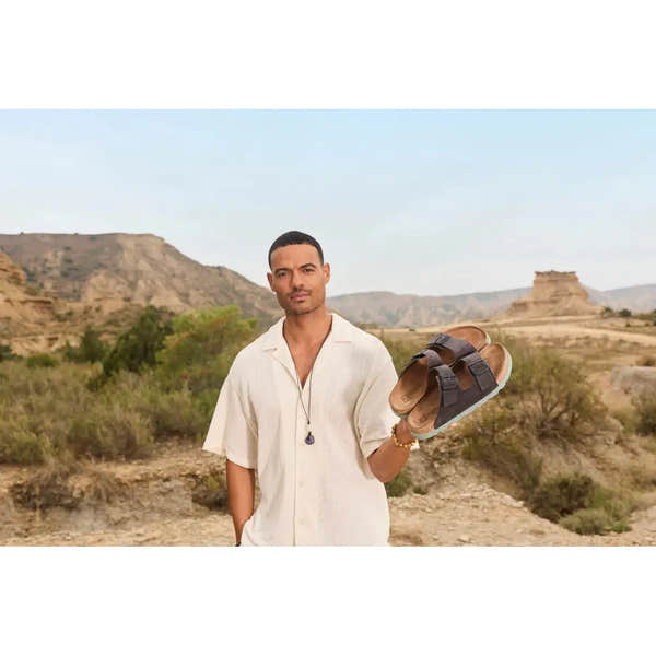 Rieker 21190-45 Men's wide fit grey suede buckle mules, casual slip-on footwear in a desert landscape setting.