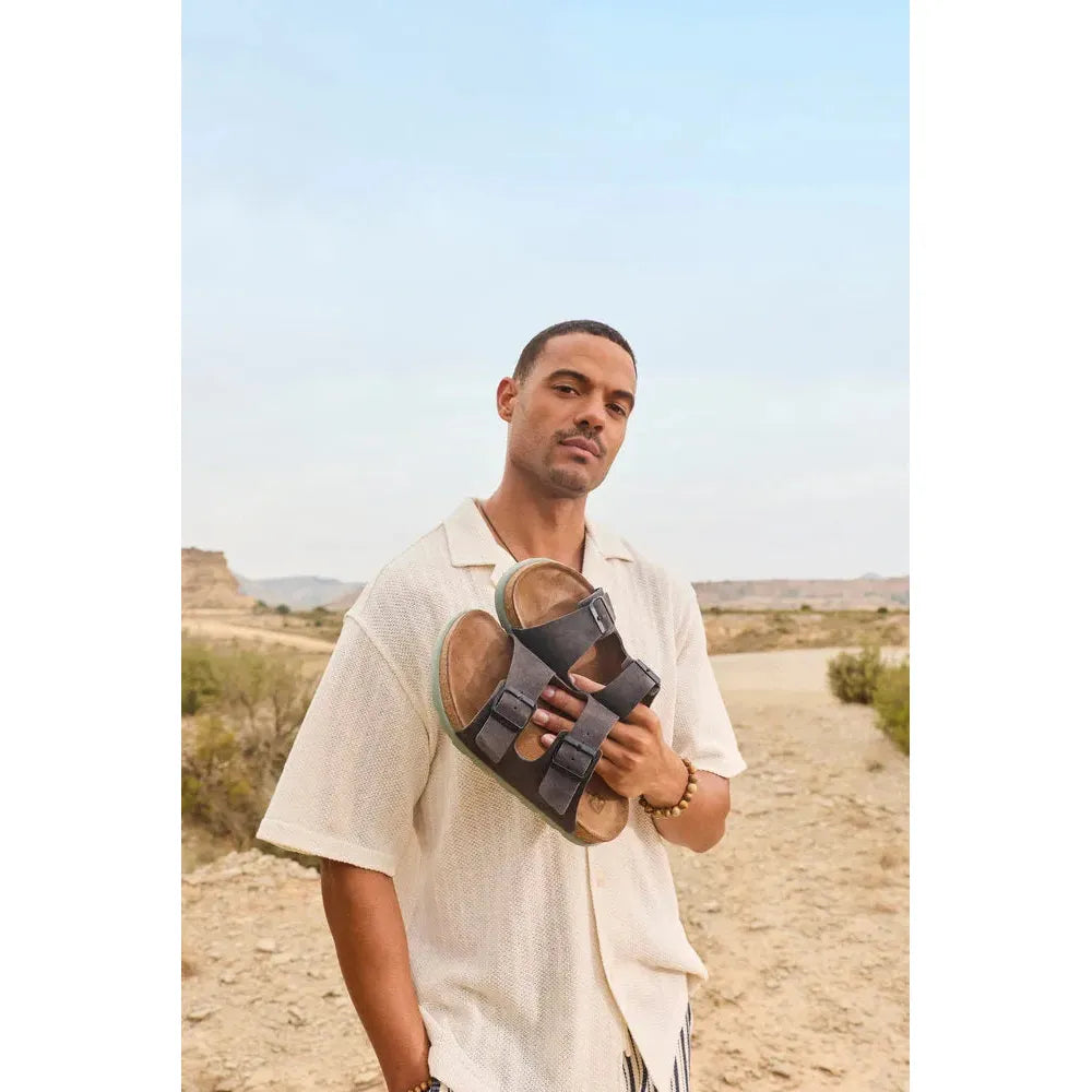 Rieker 21190-45 men's wide fit grey suede buckle mules, casual slip-on footwear with adjustable straps, in outdoor desert setting.