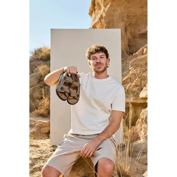 Rieker 25055-64 men's wide fit touch fastening sandals, beige with adjustable straps, outdoors in a rocky desert landscape.