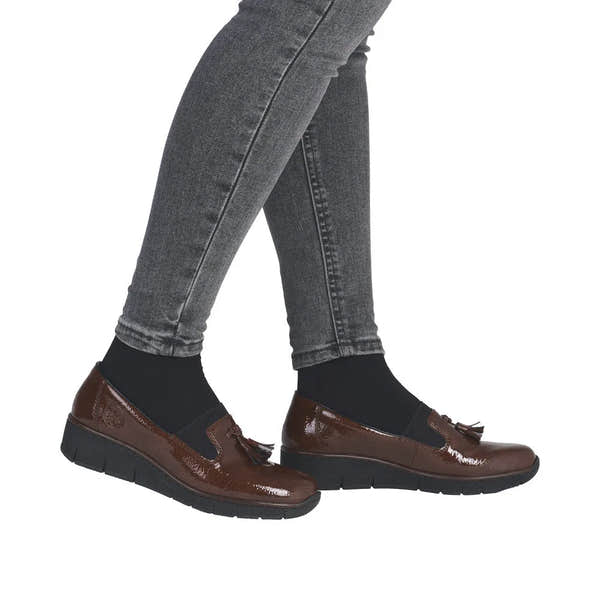 Rieker 53751-24 ladies brown slip-on shoes with tassel detail, molded rubber sole, and glossy finish, paired with black socks and gray jeans.