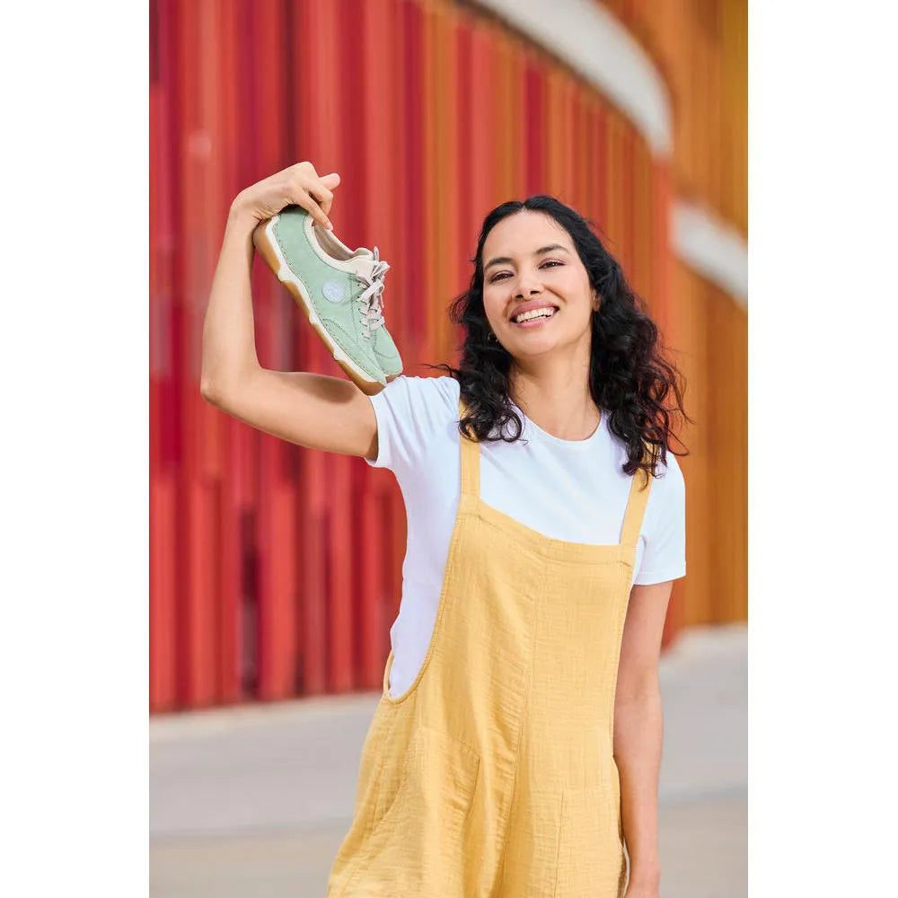 Woman holding mint lace-up shoes outdoors, wearing a white t-shirt and yellow overall, colorful background.