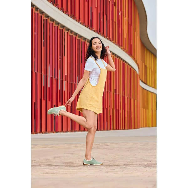 Woman in yellow dress and white t-shirt wearing mint lace-up shoes, standing in front of a colorful wall.