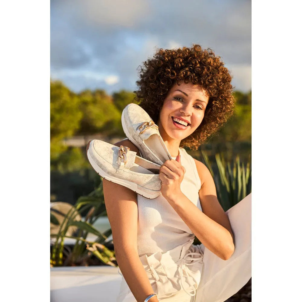 Women holding beige slip-on shoes, casual Rieker 58944-61, lightweight, comfortable beige flat sneakers with bows, outdoor setting.