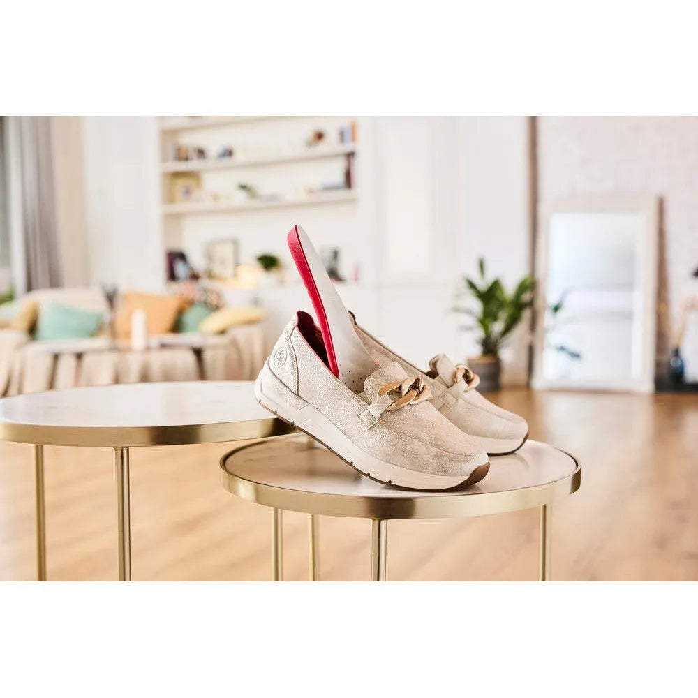 Rieker 58944-61 women's beige slip-on shoes with bow detail, placed on round tables in a bright living room setting.