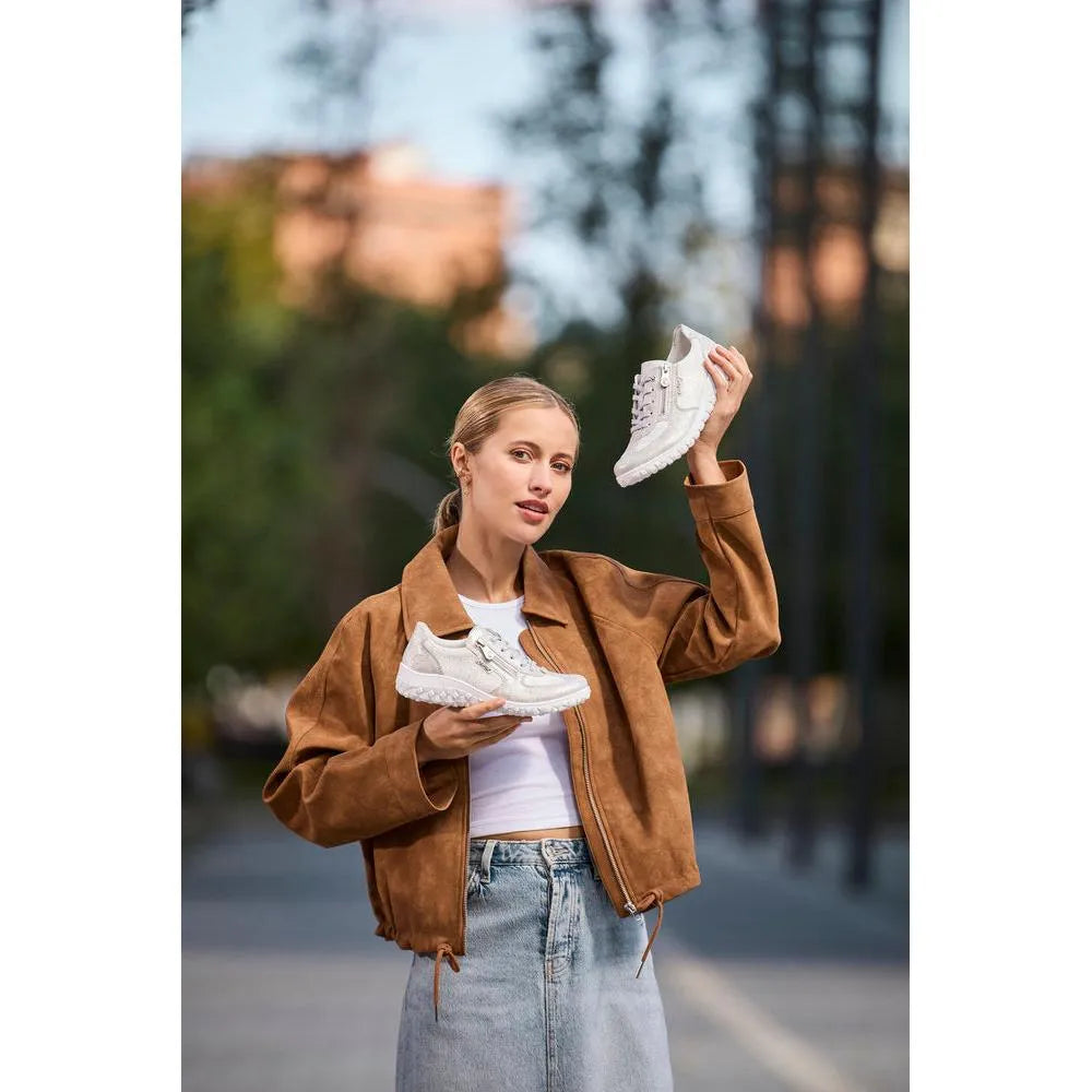 Woman holding white Rieker 59501-90 wide fit zip and lace sneakers outdoors, brown jacket, casual style.