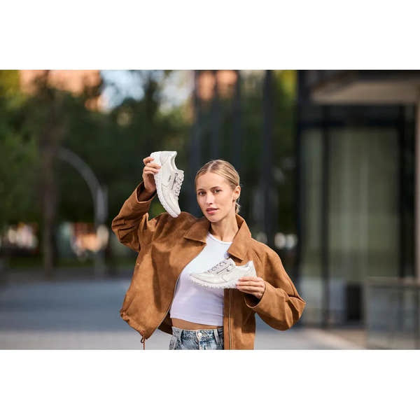 Woman holding white and silver Rieker 59501-90 wide fit sneakers outdoors, wearing a brown jacket and white crop top.