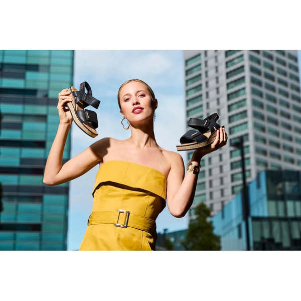 Woman holding black Rieker 63781-00 ladies touch fastening sandals outdoors in front of modern buildings.