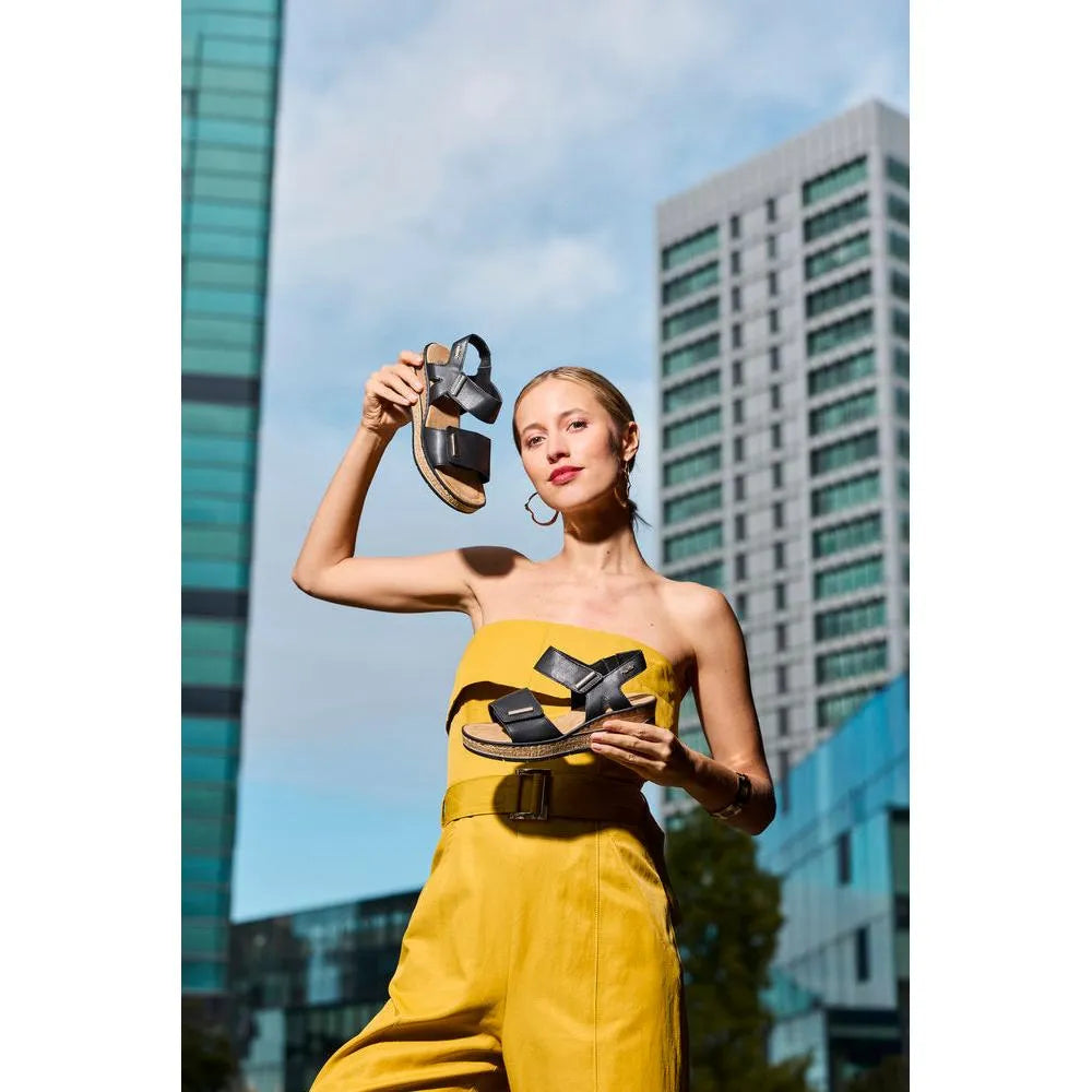 Rieker 63781-00 women's black touch fastening sandals held by woman in yellow jumpsuit, urban city background.