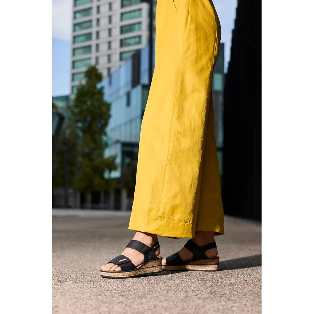 Rieker 63781-00 women's black touch-fastening sandals with wedge sole, worn by person in yellow pants, urban outdoor setting.