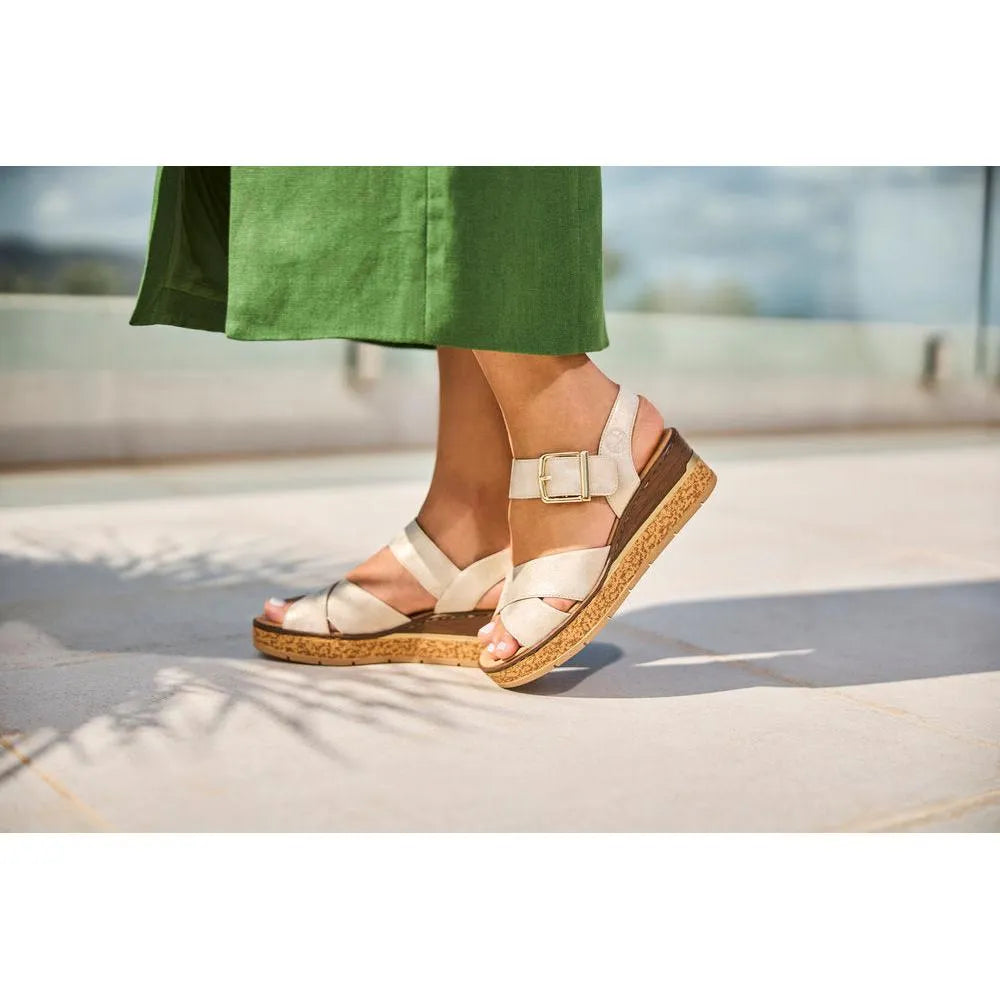 Rieker 63795-60 women's muschel buckle sandals with cream straps and cork wedge sole, worn with a green dress on a sunny outdoor terrace.