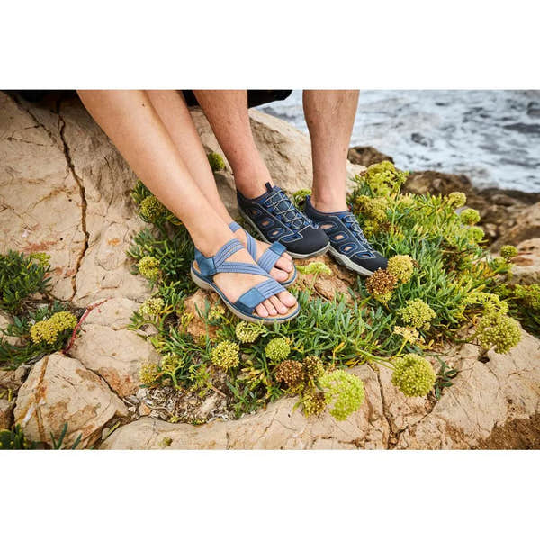 Rieker 64870-14 ladies blue Touch fastening sandals on rocky coastal terrain with green foliage.
