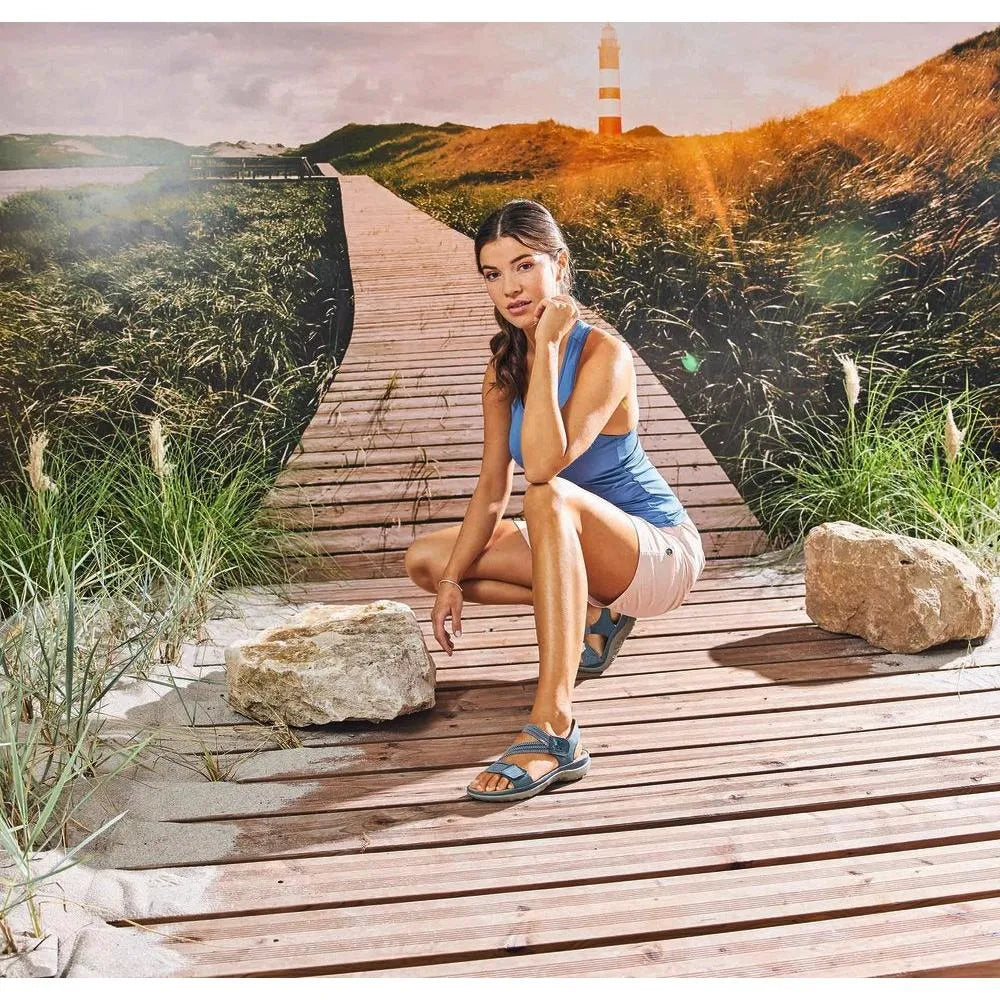 Rieker 64870-14 women's blue Touch fastening sandals on a wooden boardwalk with rocks, grass, and lighthouse background.