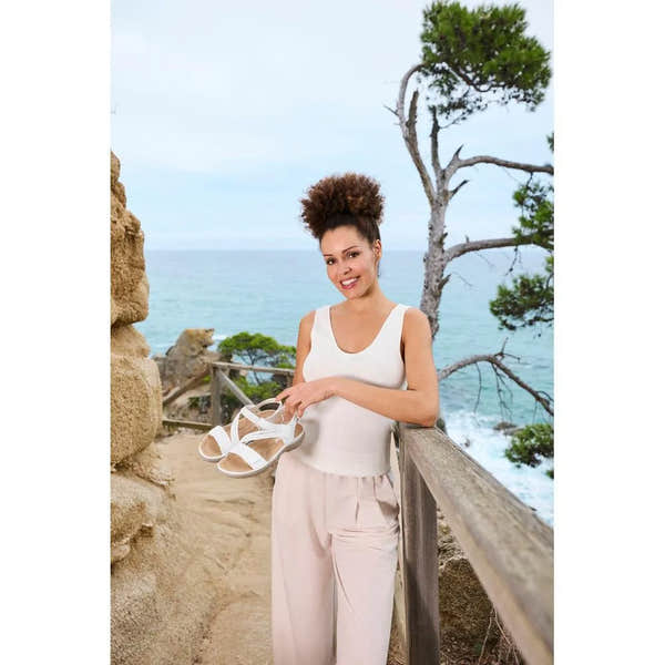 Women's white touch fastening sandals with open toe and strap detail, held by smiling woman outdoors near the coast.