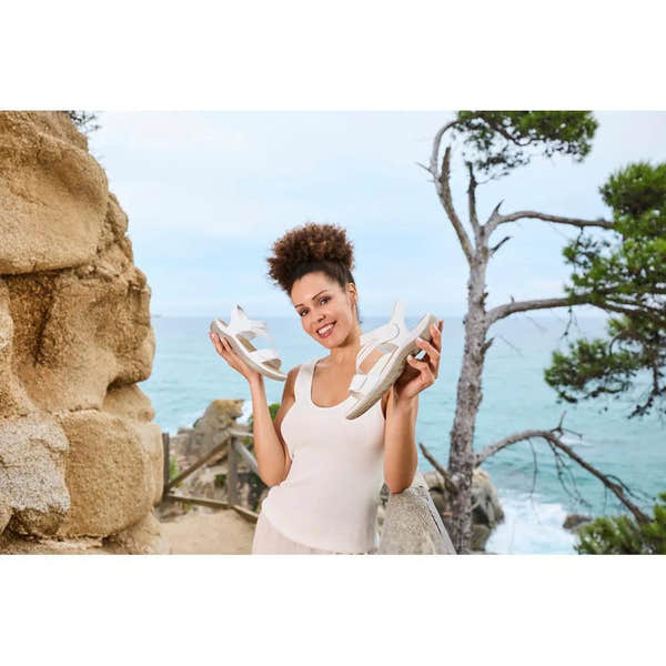 Woman holding white Rieker 64870-81 ladies sandals outdoors by the sea, with trees and rocky cliffs in the background.