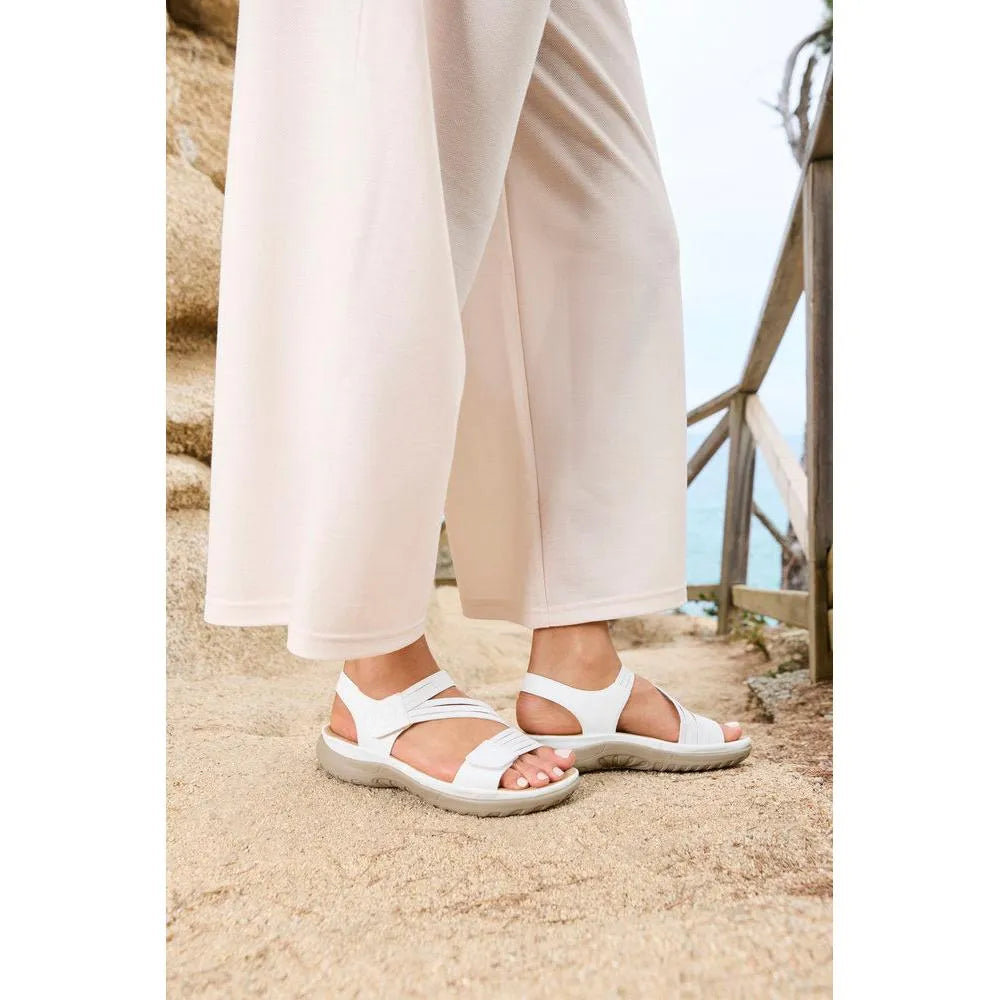 White Rieker sandals with touch fastening, featuring soft straps and a cushioned sole, paired with light beige pants.
