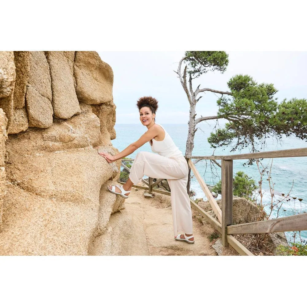 White touch fastening sandals with open toes and chunky heels worn by a woman near rocky cliffs and trees by the sea.