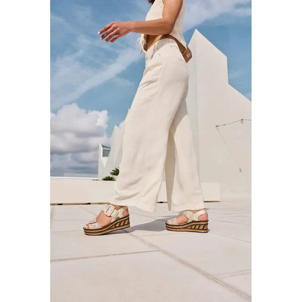Rieker 68172-60 beige touch fastening sandals on a woman with wide-leg cream pants and a blue sky background.