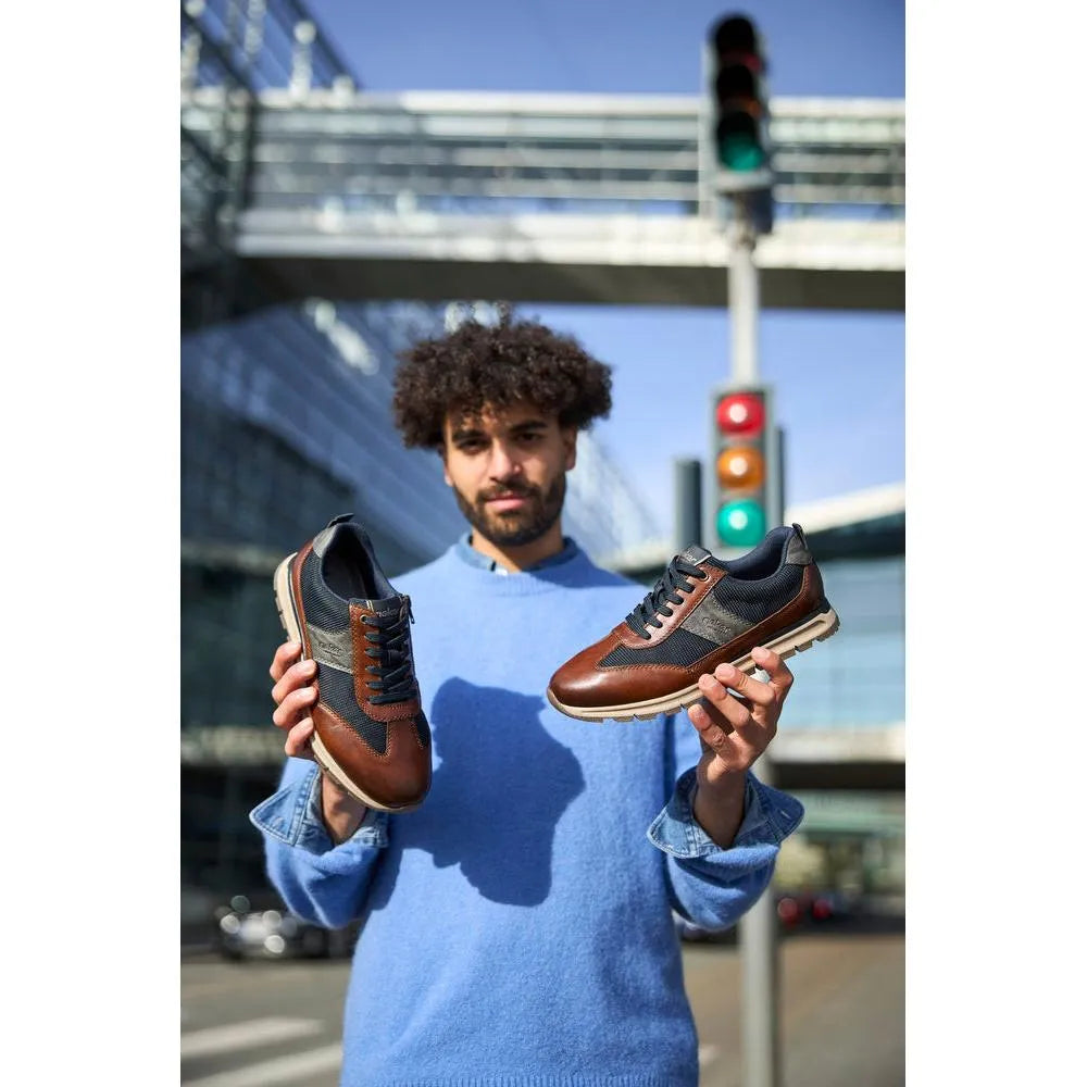 Man holding Rieker B0507-24 men's brown leather sneakers with zip and lace closure, urban background and traffic light.