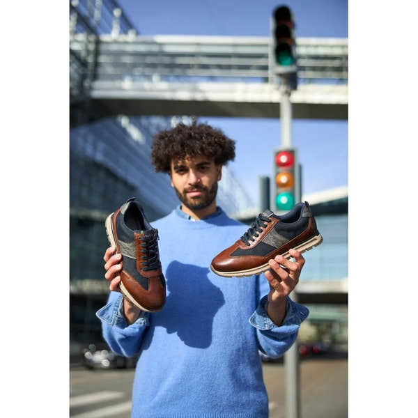 Man holding Rieker B0507-24 men's brown leather sneakers with zip and lace closure, urban background and traffic light.