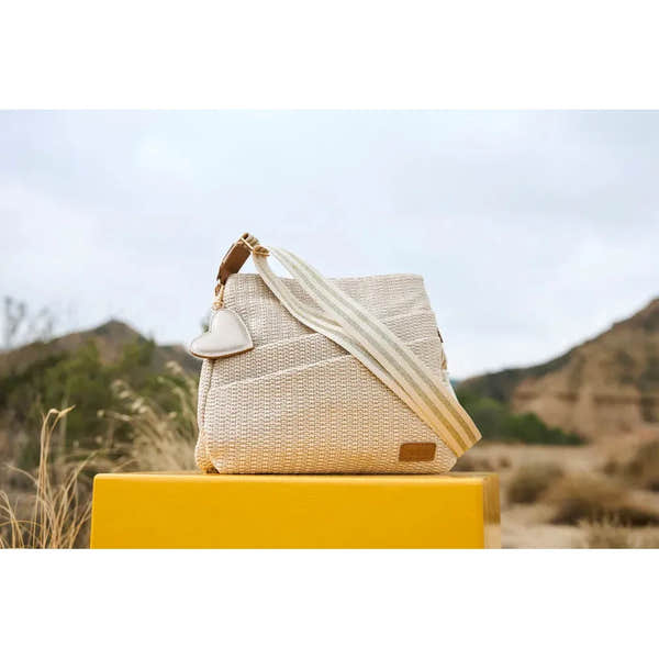 Rieker H1004-62 pale gold handbag with textured fabric, adjustable striped strap, and small heart-shaped accessory, outdoors.