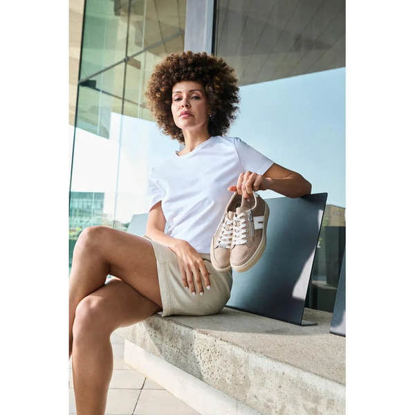 Woman holding beige suede Rieker L5903-20 shoes, sitting outdoors with modern glass building background.