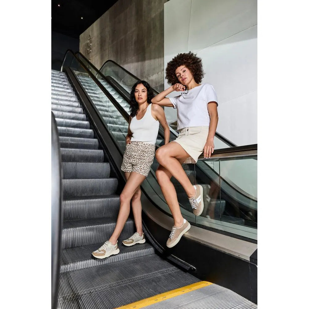 Two women in casual outfits showcase Rieker L5903-20 camel suede shoes with laces and zips, on an escalator.
